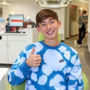 Young boy in blue cloud-patterned shirt giving thumbs up in orthodontic office.