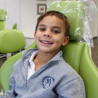 Young boy smiling in dental chair at orthodontic office with green headrest visible.