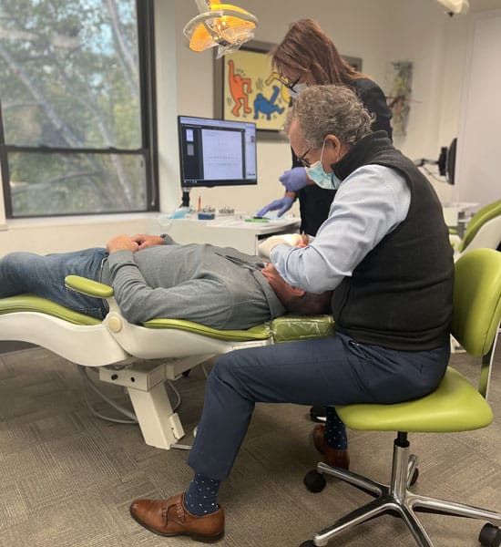 Orthodontist examining patient's teeth in modern dental office with green chair and computer monitor visible.