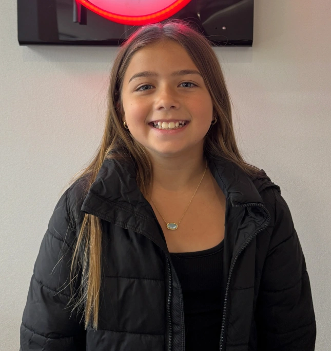 Young girl smiling at camera wearing black jacket in indoor office setting with neon sign background.