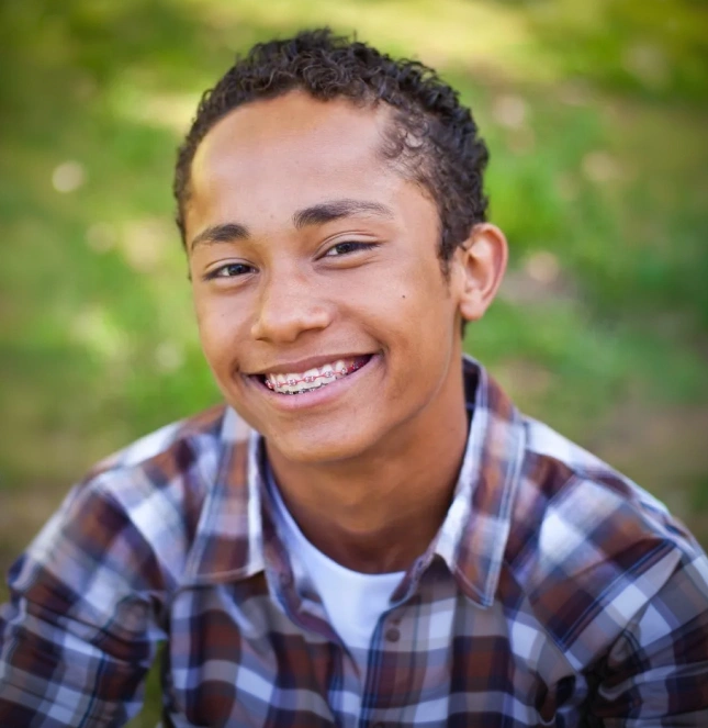 Smiling young boy wearing plaid shirt against blurred green outdoor background.