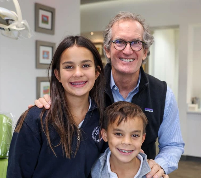 Man and two children smiling together in orthodontic office setting with framed artwork on walls.